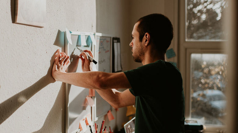 A young man, who is clearly organised in nature, writes reminders and affirmations on a white board on his wall, within a small home office environment.