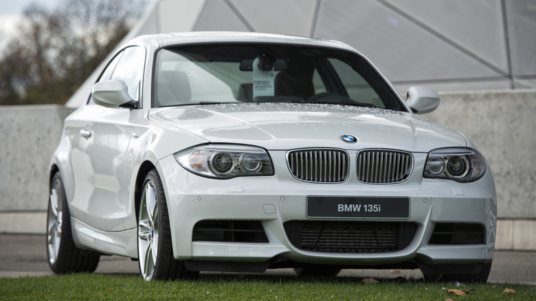 A 2013 E82 BMW 135i finished in white, parked on grass in front of a grey building
