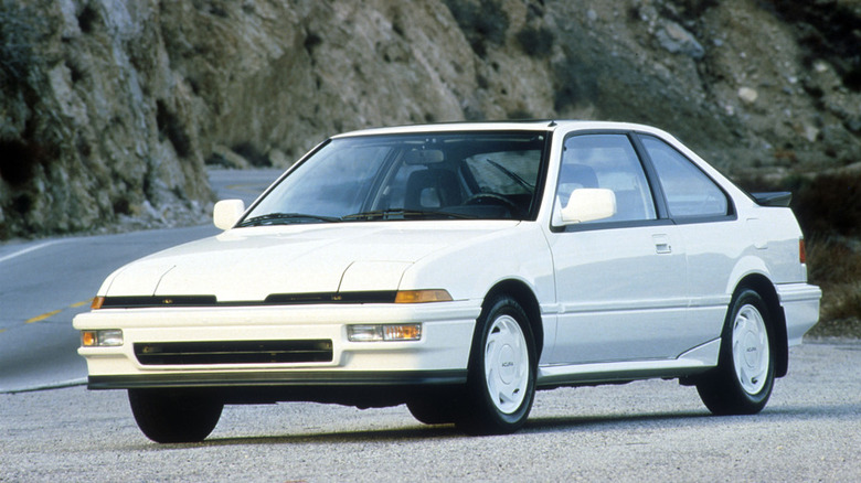 A white first-generation Acura Integra two-door coupe parked on a mountain road, showing its clean 1980s wedge-shaped design.