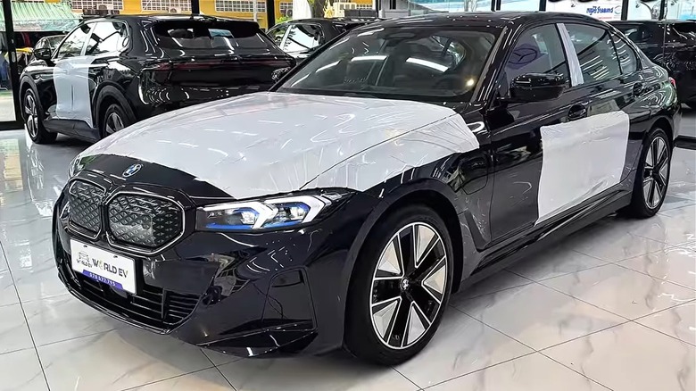 A black 2026 BMW i3 sedan sitting pretty in a busy showroom partially wrapped in white protective film on the hood and sides while being surrounded by other luxury EVs in the background.