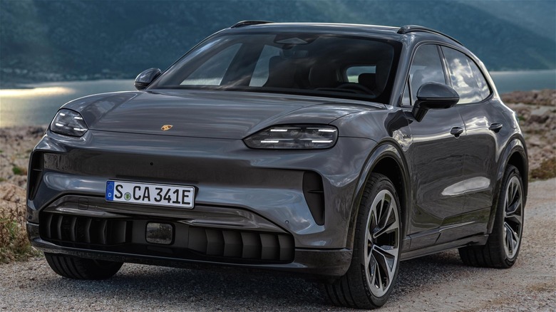 A sleek gray Porsche Cayenne EV is parked on a gravel road overlooking a calm lake and rugged mountains under moody clouds.
