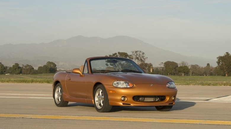 A bronze Mazda MX-5 Miata convertible (NB generation) parked on a runway with mountains in the background.
