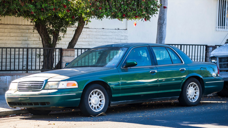 A green Ford Crown Victoria sedan is parked on the street under a tree.