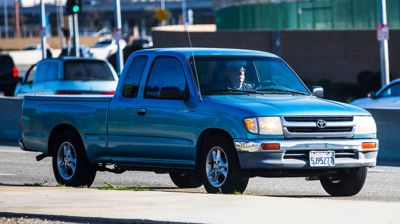 A blue first generation Toyota Tacoma parked on the side of the road, with a older gentleman in the driver's seat.