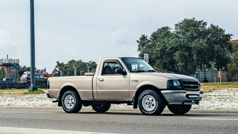 A beige Ford Ranger driving down a city street, with trees and a building in the background.
