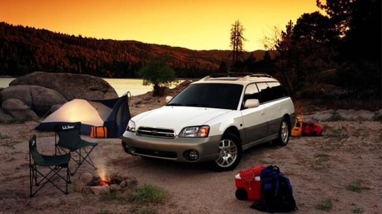 A two-tone Subaru Outback wagon from the early 2000s parked at a campsite near a lake.