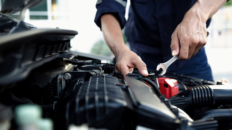 A man wearing glasses and a green t-shirt works under the hood of a car, using a screwdriver to make adjustments to the engine.