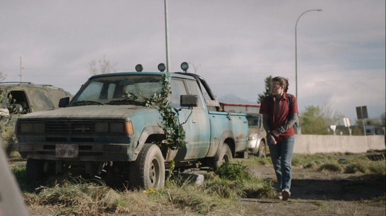 Ellie walks past an overgrown Datsun 720 pickup truck on a deserted roadside filled with abandoned vehicles in "The Last of Us."