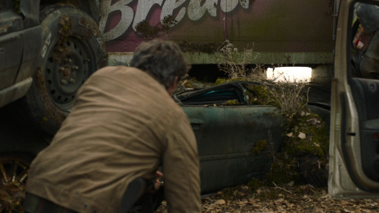 Joel crouches near a moss-covered, overturned Ford Taurus wedged under a trailer in a tense scene from "The Last of Us."