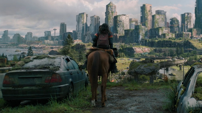 Ellie and Dina approach the moss-covered ruins of Seattle on horseback, passing a decaying BMW 3 Series sedan and other overgrown wrecks in "The Last of Us."
