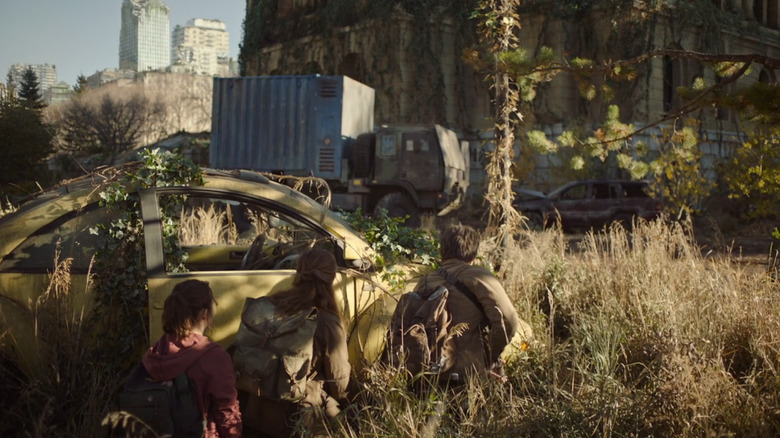 Joel, Tess, and Ellie crouch behind a vine-covered yellow Volkswagen New Beetle as they scout the overgrown ruins of the city in "The Last of Us."