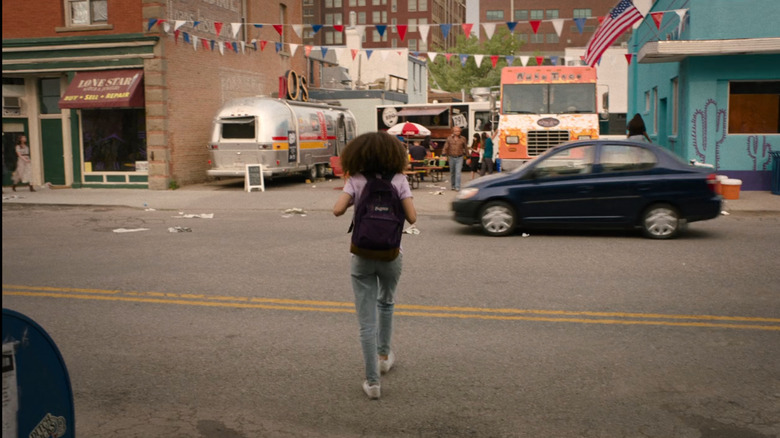 A navy blue Toyota Echo drives past as a Joel's daughter, Sarah,runs toward a bustling food truck scene in "The Last of Us."