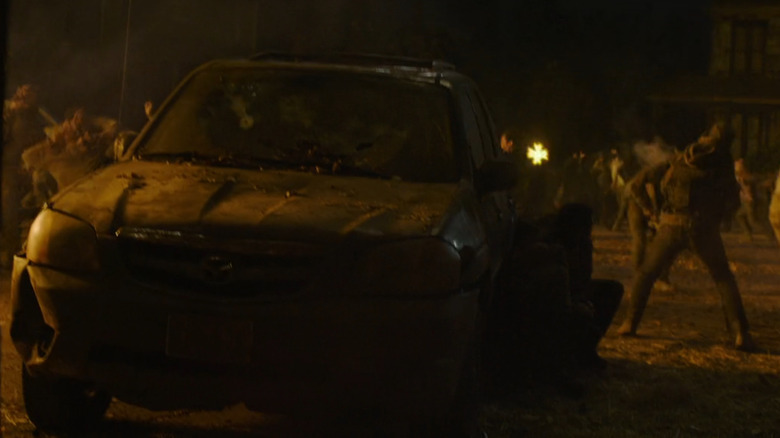 A battered Mazda Tribute sits in the foreground as chaos erupts behind it during a nighttime infected attack in "The Last of Us."