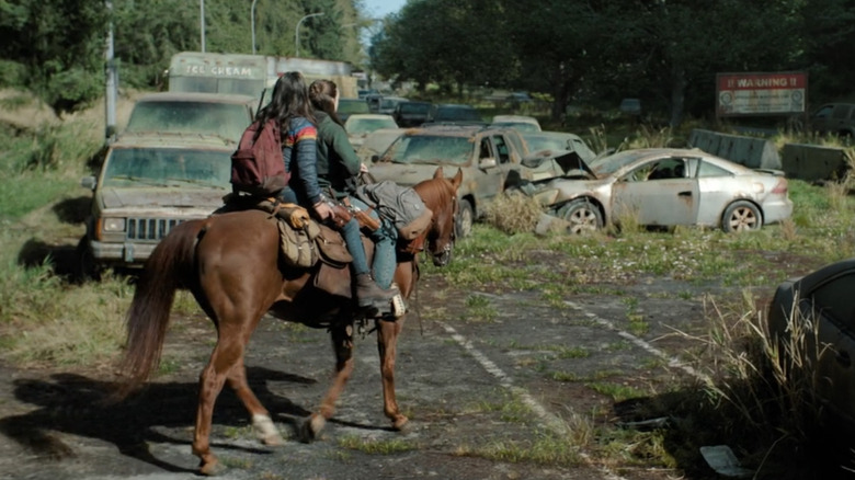 Ellie and Dina ride past a rusting silver Honda Accord coupe among a pileup of abandoned cars on an overgrown highway in "The Last of Us."