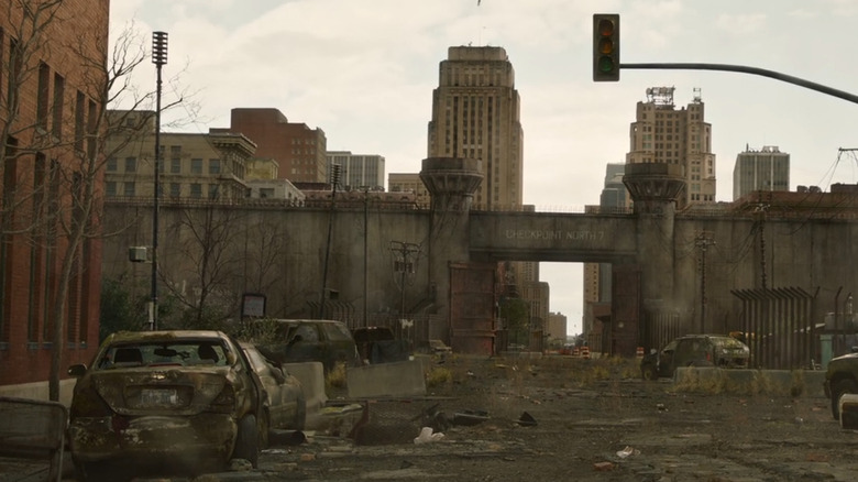 A decaying Chevrolet Epica sits abandoned outside Checkpoint North 7, framed by the crumbling ruins of Kansas City's quarantine zone in "The Last of Us."