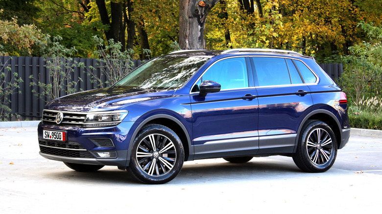 A 2020 Volkswagen Tiguan in a dark colorway parked in a lot with trees in the background
