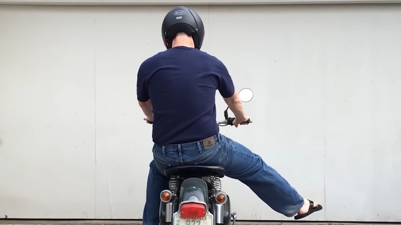 A biker wearing a blue t shhirt and a helmet sticking his right leg out on a parked bike