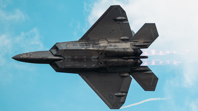 The underside of an F-22 Raptor with afterburner thrust visible as it zooms under a cloudy sky