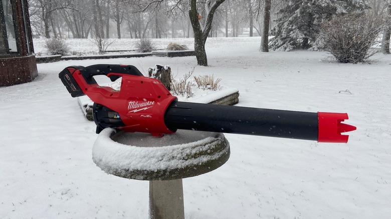 A Milwaukee M18 Fuel leaf blower with a red velocity nozzle attachment sits on a snow-covered bird bath outdoors.