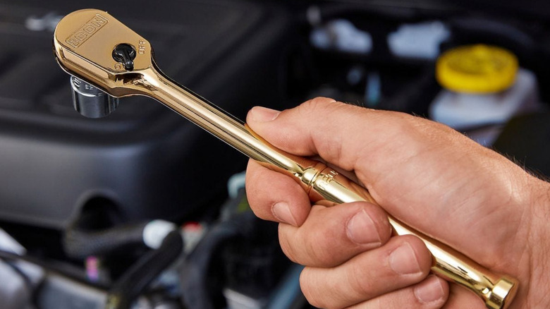 A hand holding a gold-plated ratchet, vehicle engine in background.