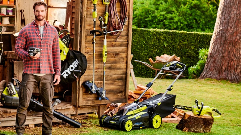 A man stands in front of a shed full of Ryobi tools, holding a Ryobi battery
