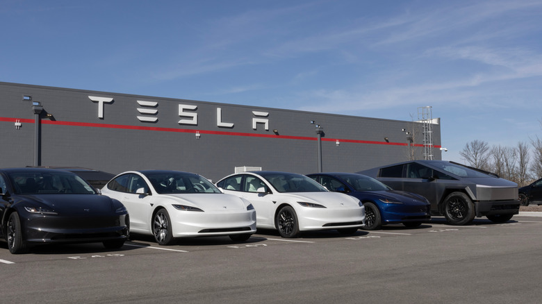 Tesla vehicles parked in a lot outside, with a tesla building visible in the background