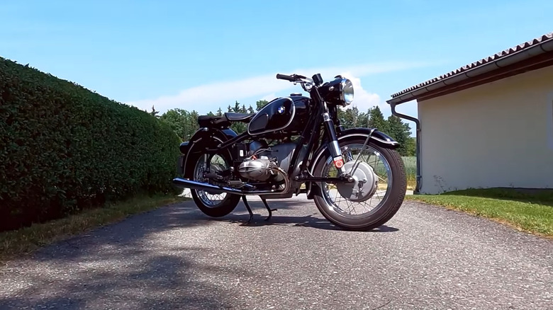 A navy and silver BMW R69S standing on a gravel road between a hedge and a shed.