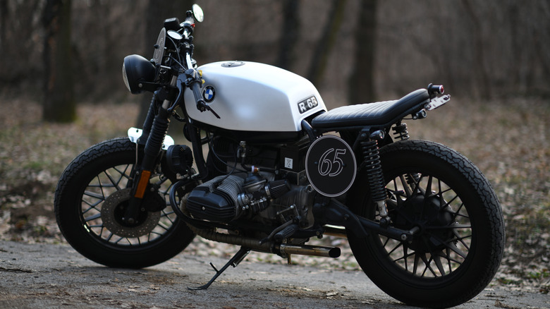A white BMW R65 Cafe Race motorbike standing in a wooded area with trees in the background