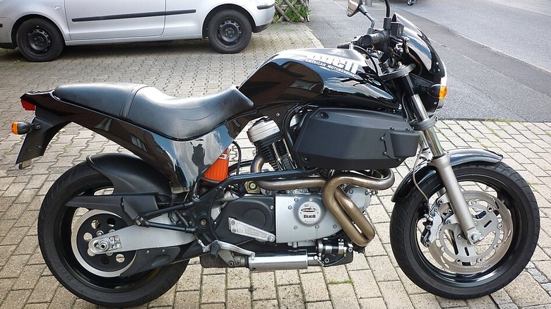 A black Buell M2 Cyclone sitting on paved brick, outside in a parking lot with a silver car in the background
