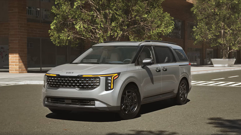 A gray Kia Carnival driving through a small town road.