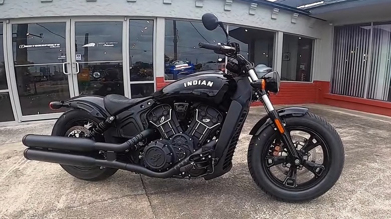 A black Indian Scout Bobber Sixty motorcycle parked in front of a modern dealership building with large glass windows and a red-brick lower facade.