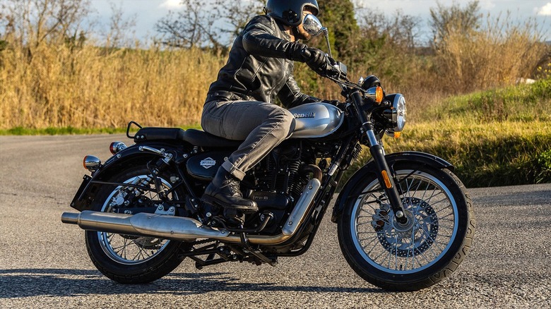 A rider in a black helmet and jacket operating a black Benelli Imperiale 400 motorcycle on a curved rural road, with grassy fields and trees in the background under a partly cloudy sky.