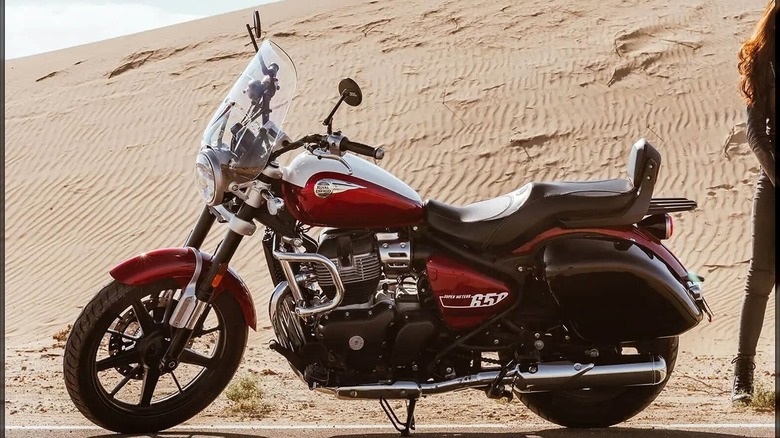 A Royal Enfield Super Meteor 650 cruiser motorcycle in a two-tone red and silver color scheme, parked on the roadside with sand dunes in the background.