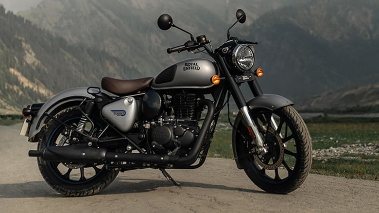 A Royal Enfield Classic 350 motorcycle with a matte finish, parked on a scenic dirt road against a mountain and cloud backdrop.