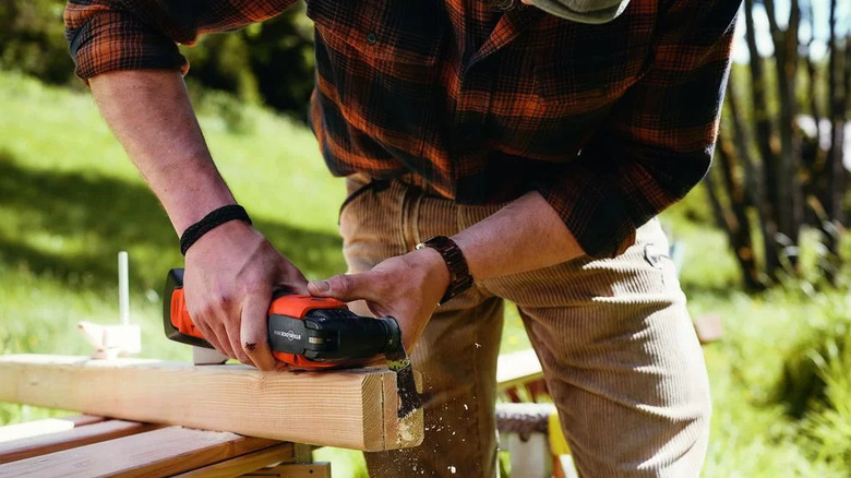 A person in a plaid shirt using a Fein cordless multi-tool to cut a wooden board outdoors.
