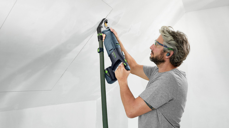 A person using a Festool Vecturo cordless oscillating multi-tool with dust extraction to cut into a ceiling.