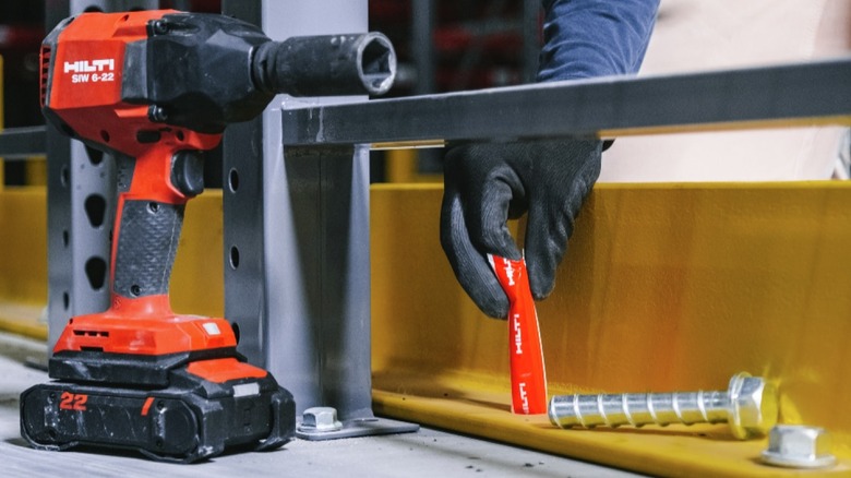 A worker using a Hilti impact wrench and red Hilti torque tool to install a concrete anchor bolt into a yellow steel beam.