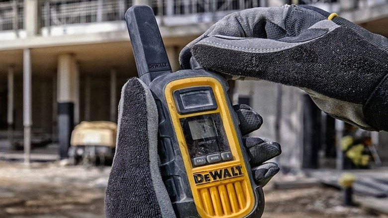 Close-up of gloved hands holding a rugged yellow-and-black DeWalt 2-Watt Heavy Duty Walkie-Talkie at a construction site.