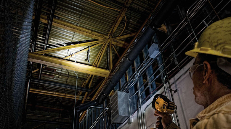 A construction worker shines a DeWalt cordless flashlight upward to inspect an industrial ceiling.