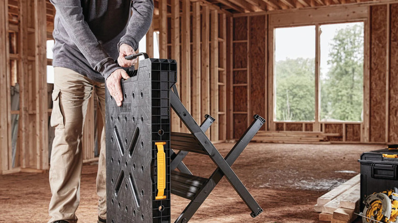 A person setting up a black DeWalt Express Folding Workbench inside a partially finished building.