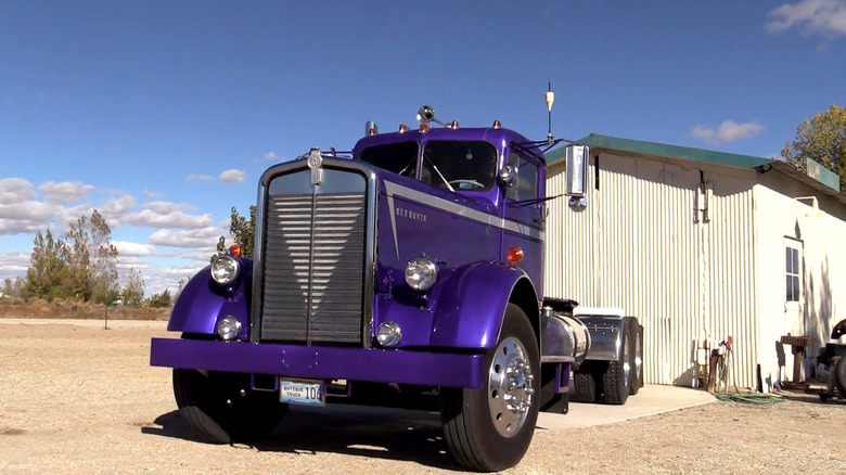 A beautiful purple 1956 Kenworth 825 series cabover truck parked outside a garage