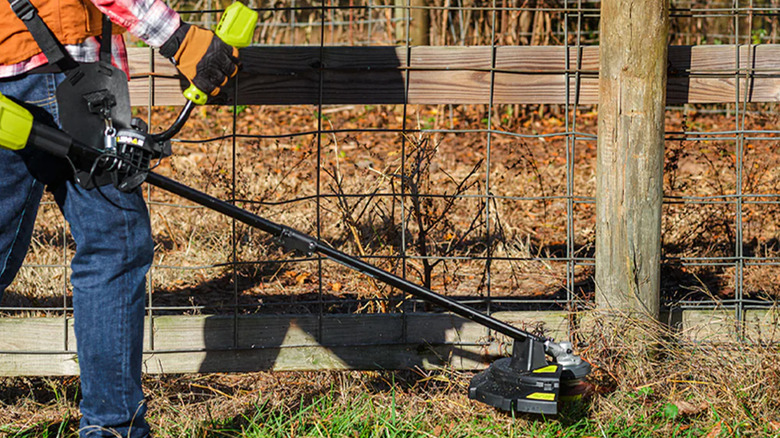 A person using a brush cutter to clear weeds by a fence.