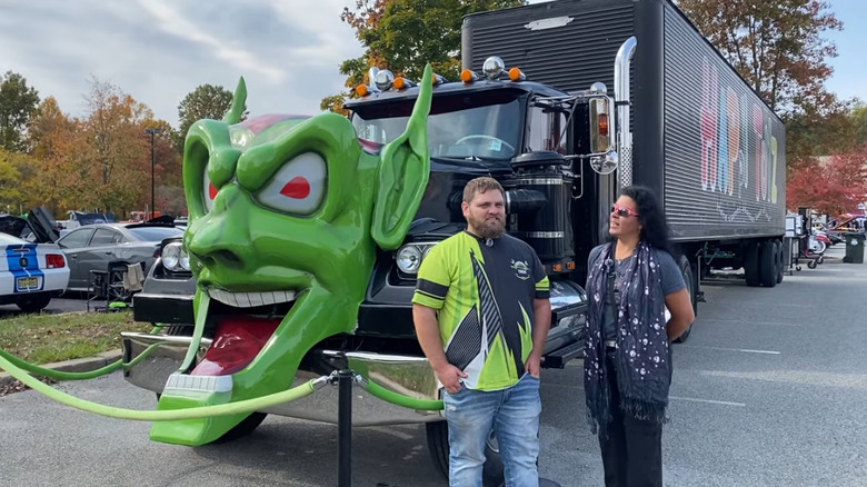 A black semi truck with a large green goblin face on the front is displayed at a car show, with two people standing in front of it.