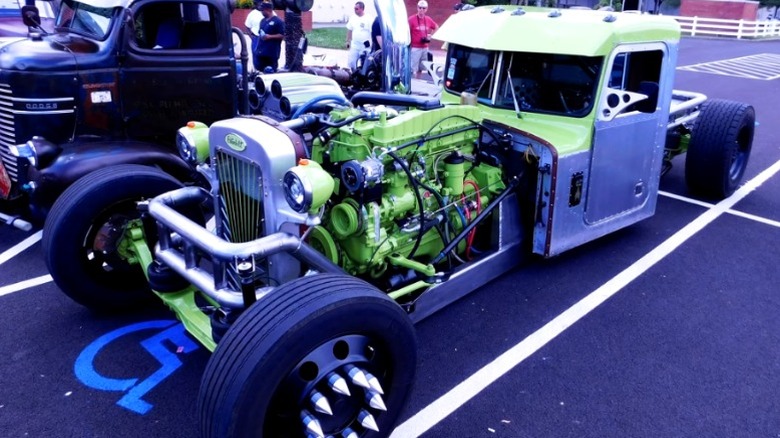 A custom rat rod semi truck with a bright green exposed engine, spiked wheels, and a silver and green cab.