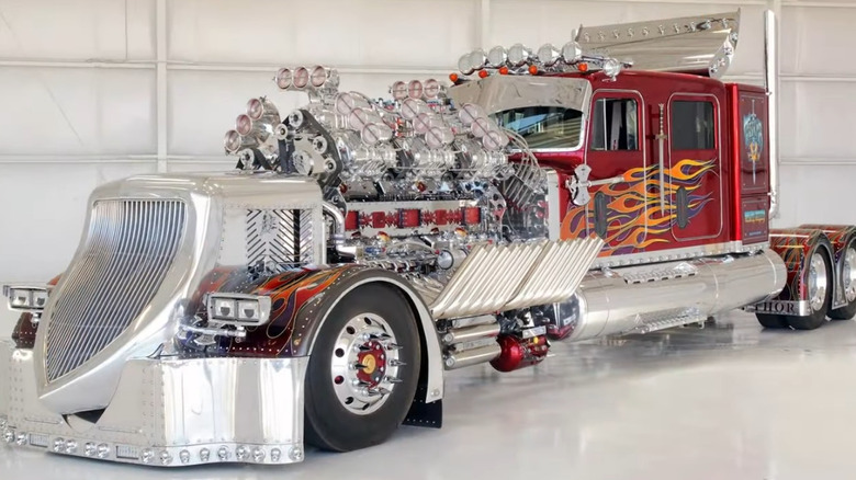 An extreme custom semi truck with a polished chrome front end, massive exposed multi-engine setup with multiple superchargers, and a red cab decorated with flame graphics.
