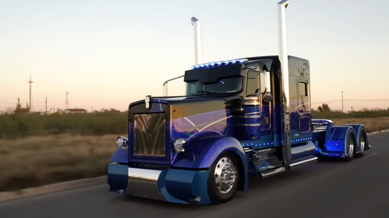 A custom deep blue semi truck with chrome exhaust stacks, glowing blue lights, and a sleek modern design drives down a road at sunset.