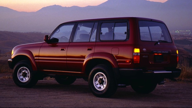 A 1992 red Toyota Land Cruiser FJ80 001 sits parked in an open, deserted-looking area, with distant lights casting a soft glow across the barren background