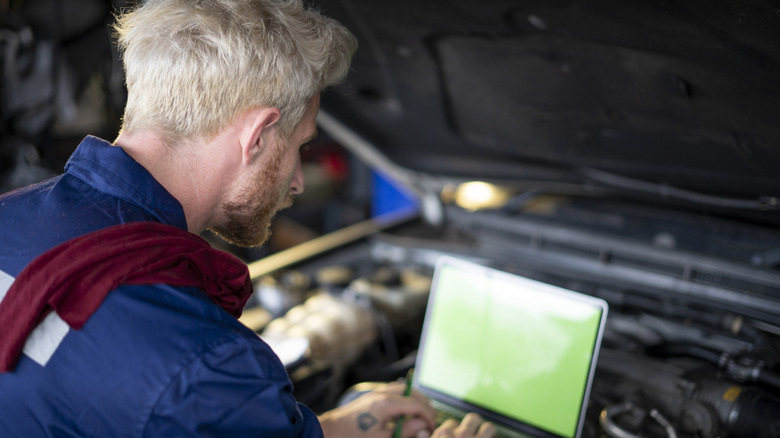 A mechanic using a laptop computer to tune a vehicle under an open hood.