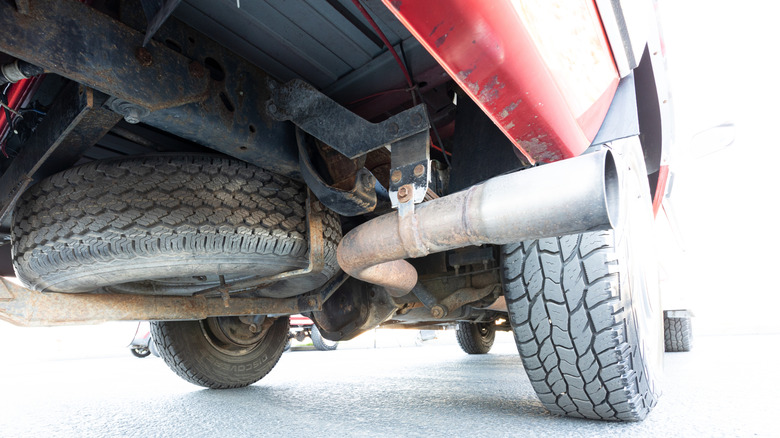 Underside view of a pickup truck showing the spare tire mounted under the bed, rear axle, leaf spring suspension, and a large aftermarket exhaust pipe.