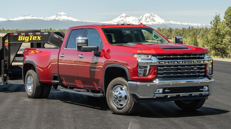A red Chevrolet Silverado 3500HD pickup truck is parked on a paved surface with mountains and trees in the background, towing a trailer with a bulldozer behind it.